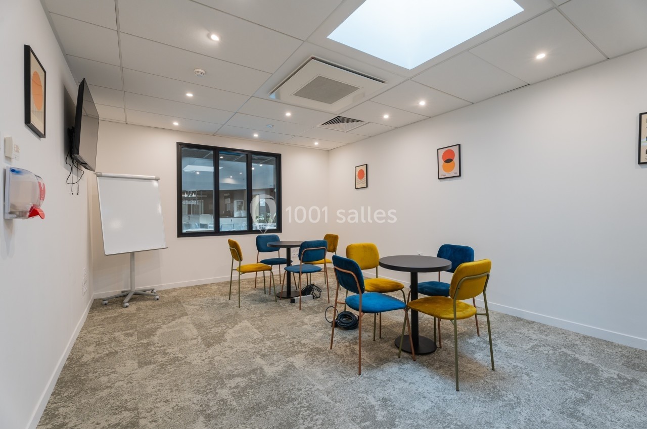 Salle de réunion lumineuse avec tables rondes, chaises colorées, tableau blanc et fenêtre donnant sur un autre espace.