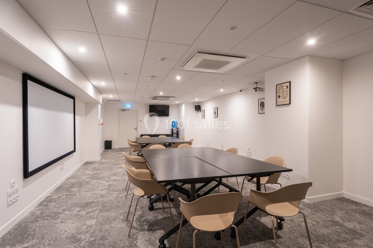 Salle de réunion moderne avec tables noires, chaises beige, écran mural et éclairage encastré.