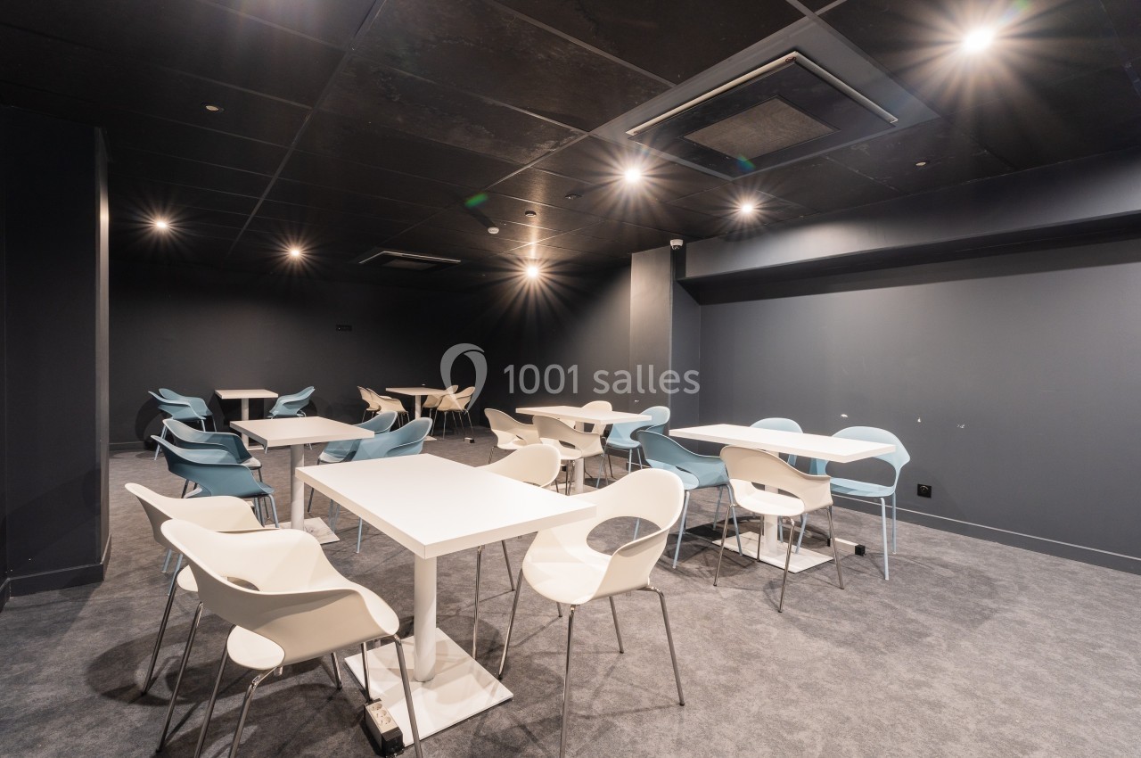 Salle moderne avec tables blanches et chaises design, éclairée par des spots au plafond sombre.