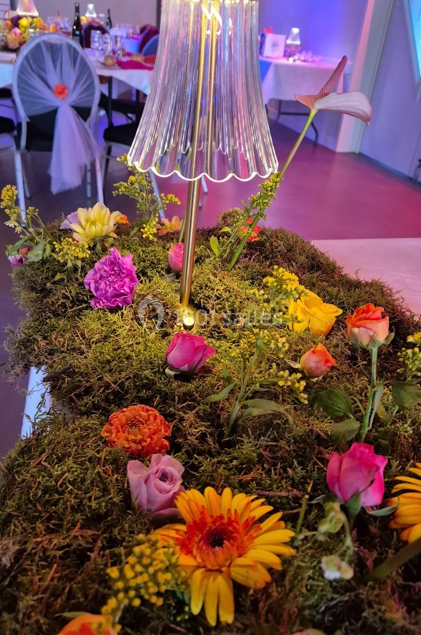 Lampe à abat-jour en verre posée sur un centre de table décoré de mousse, fleurs colorées et végétation.