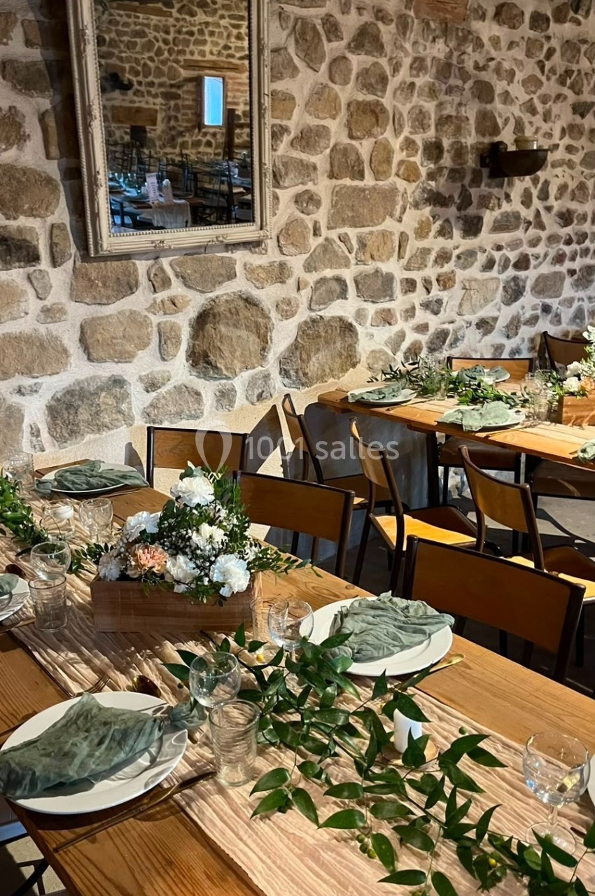 Salle rustique avec murs en pierre, tables en bois décorées de nappes vertes, fleurs et feuillages pour un repas.