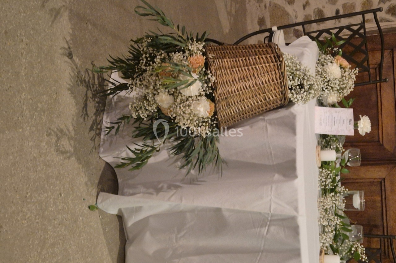 Décoration de table avec panier en osier, fleurs blanches et feuillage, disposée dans une salle au sol en pierre.