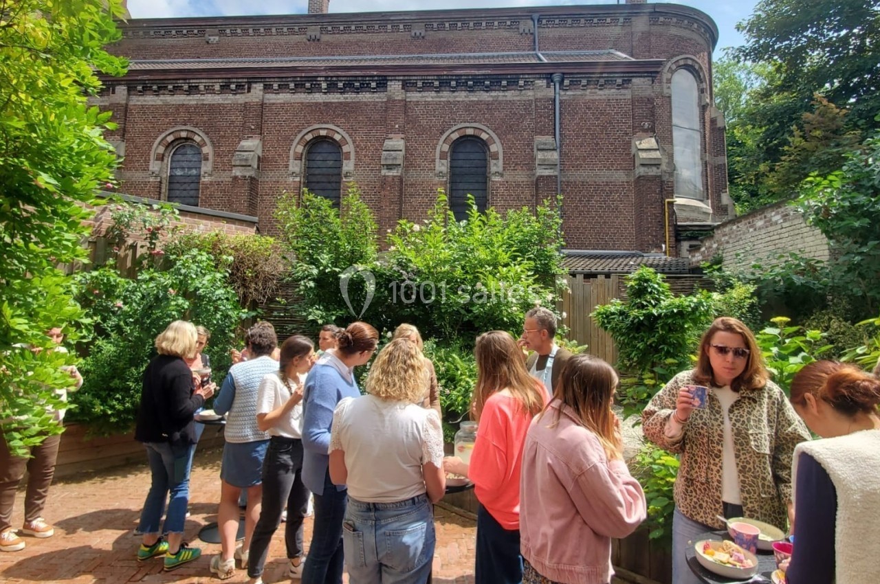 Groupe de personnes discutant et partageant un repas dans une cour extérieure bordée de verdure, près d'un bâtiment en…