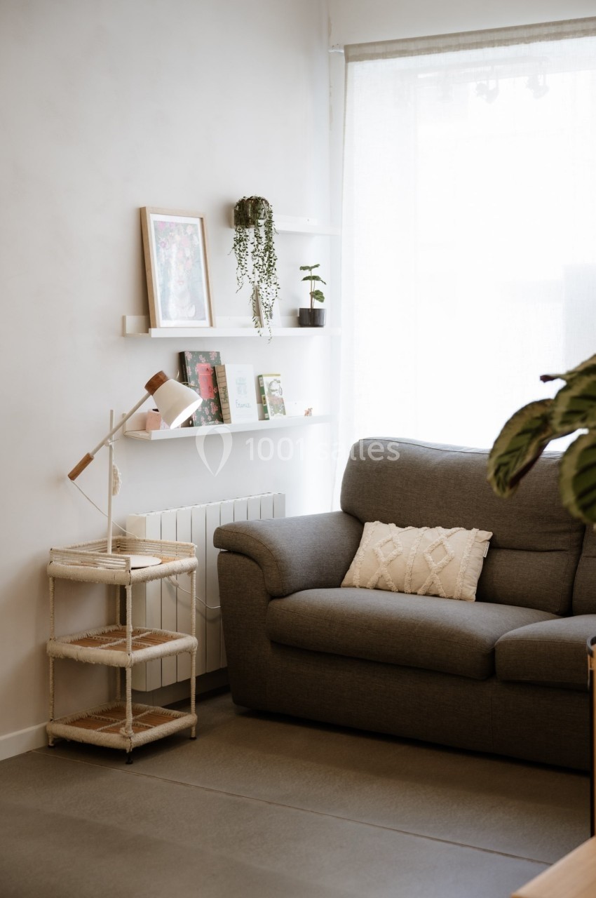 Salon lumineux avec canapé gris, étagères murales décorées, lampe sur table et grande fenêtre avec rideau clair.