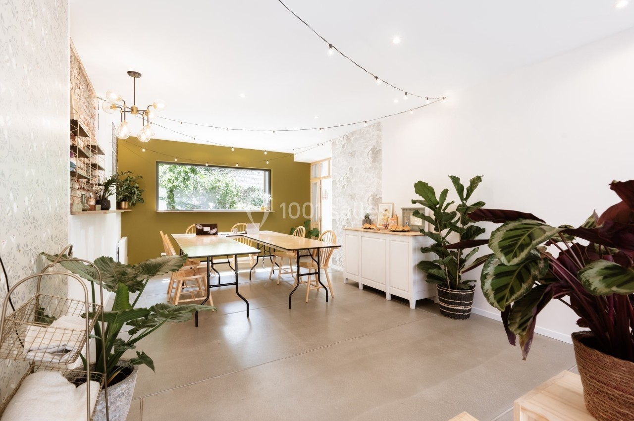 Salle lumineuse avec tables en bois, plantes vertes, murs clairs et guirlandes lumineuses suspendues.