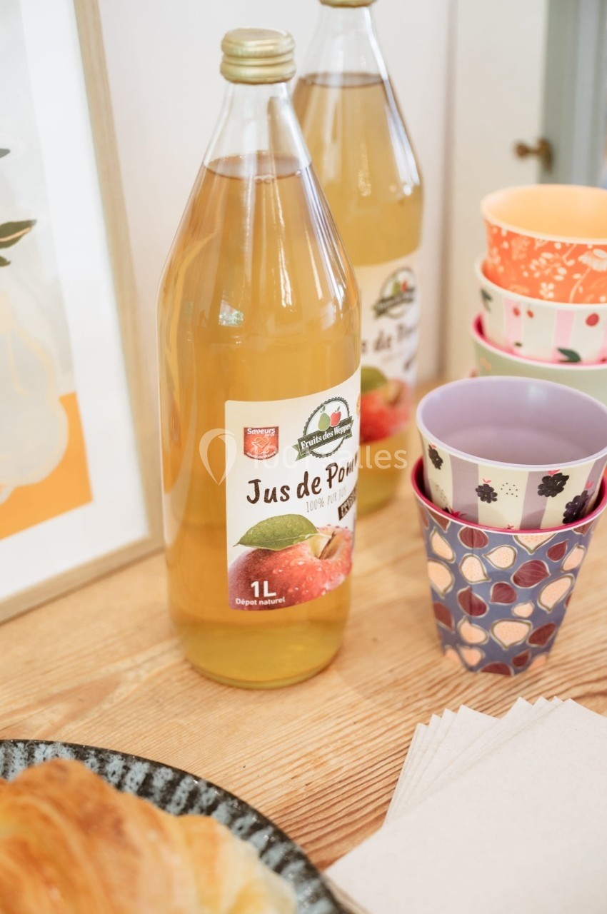 Deux bouteilles de jus de pomme sur une table en bois, entourées de gobelets colorés et d'une assiette de viennoiseries.