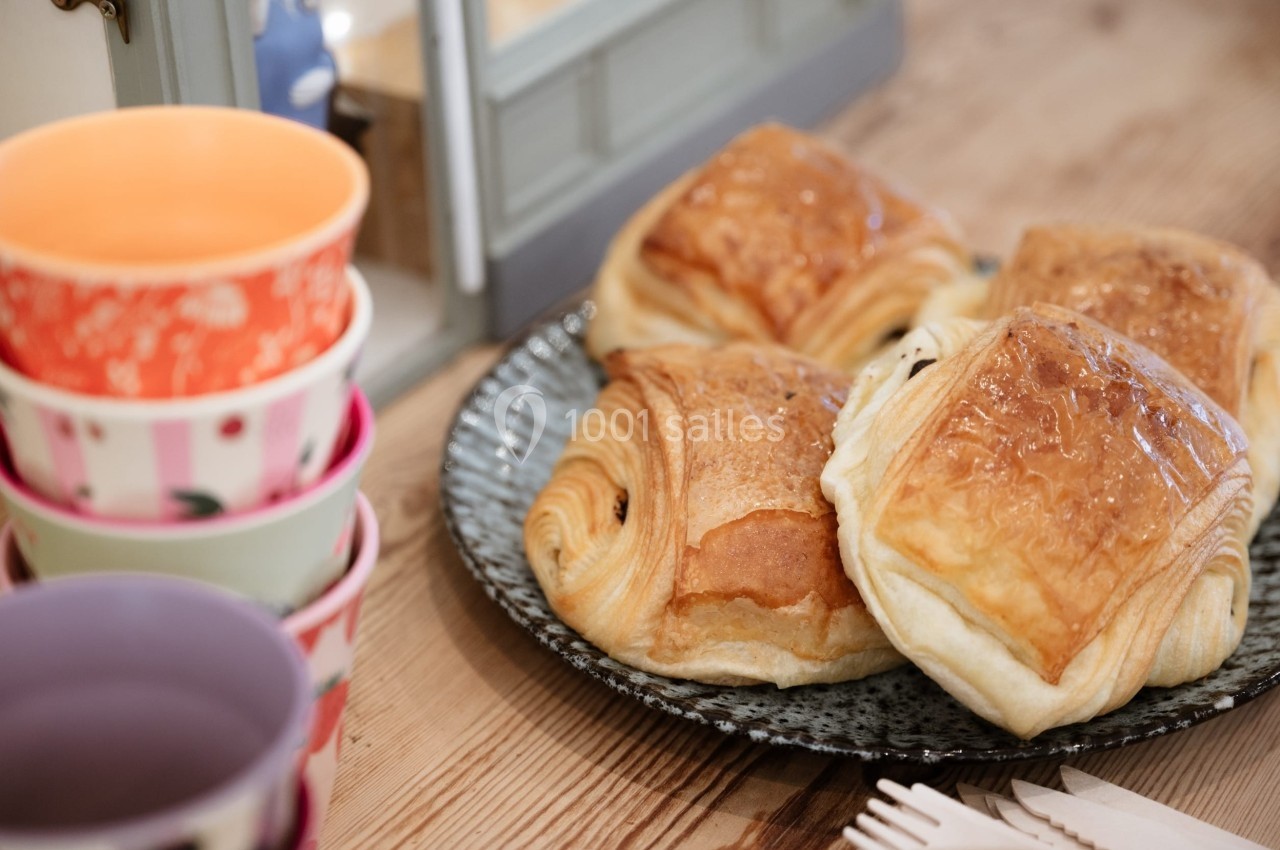 Quatre pains au chocolat dorés sur une assiette, avec des tasses colorées empilées à côté sur une table en bois.