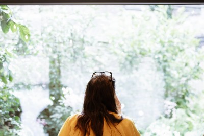 Miniature Location salle Phalempin (Nord) - L'Étoilerie #16 Une femme de dos, portant une chemise jaune, regarde par une grande fenêtre donnant sur un jardin verdoyant.