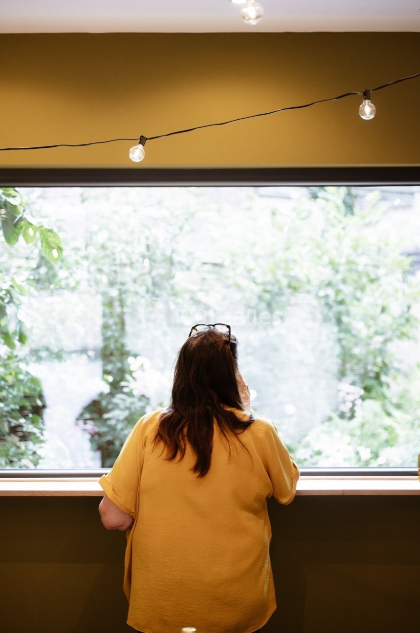 Une femme de dos, portant une chemise jaune, regarde par une grande fenêtre donnant sur un jardin verdoyant.