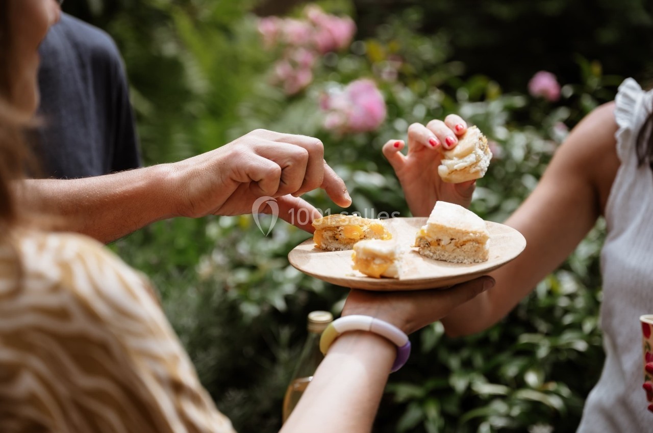 Des mains partagent des amuse-bouches sur une assiette en bois dans un jardin verdoyant.