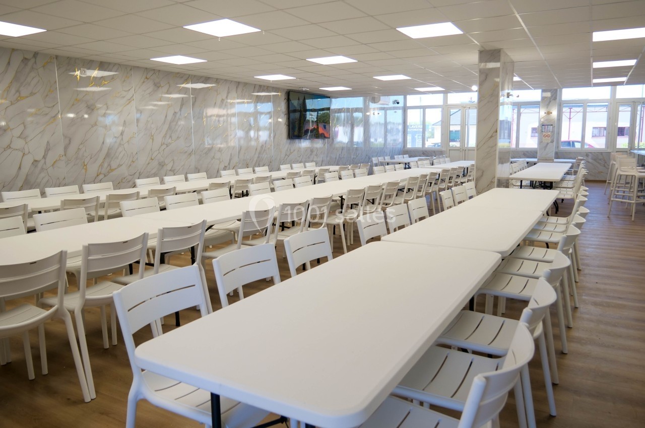 Salle lumineuse avec de longues tables blanches alignées et des chaises, sol en bois clair et murs effet marbre.