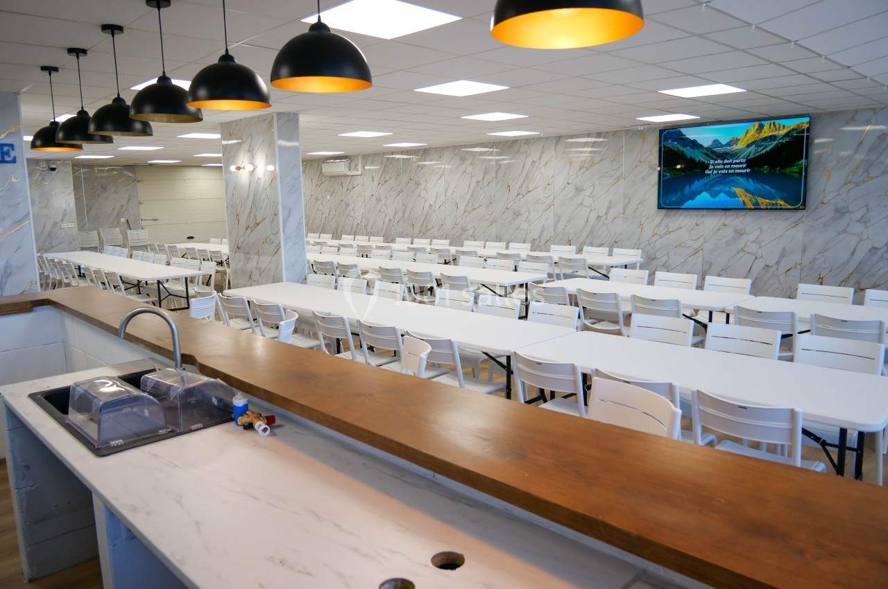 Salle de restauration lumineuse avec tables blanches alignées, comptoir en bois au premier plan et écran mural.