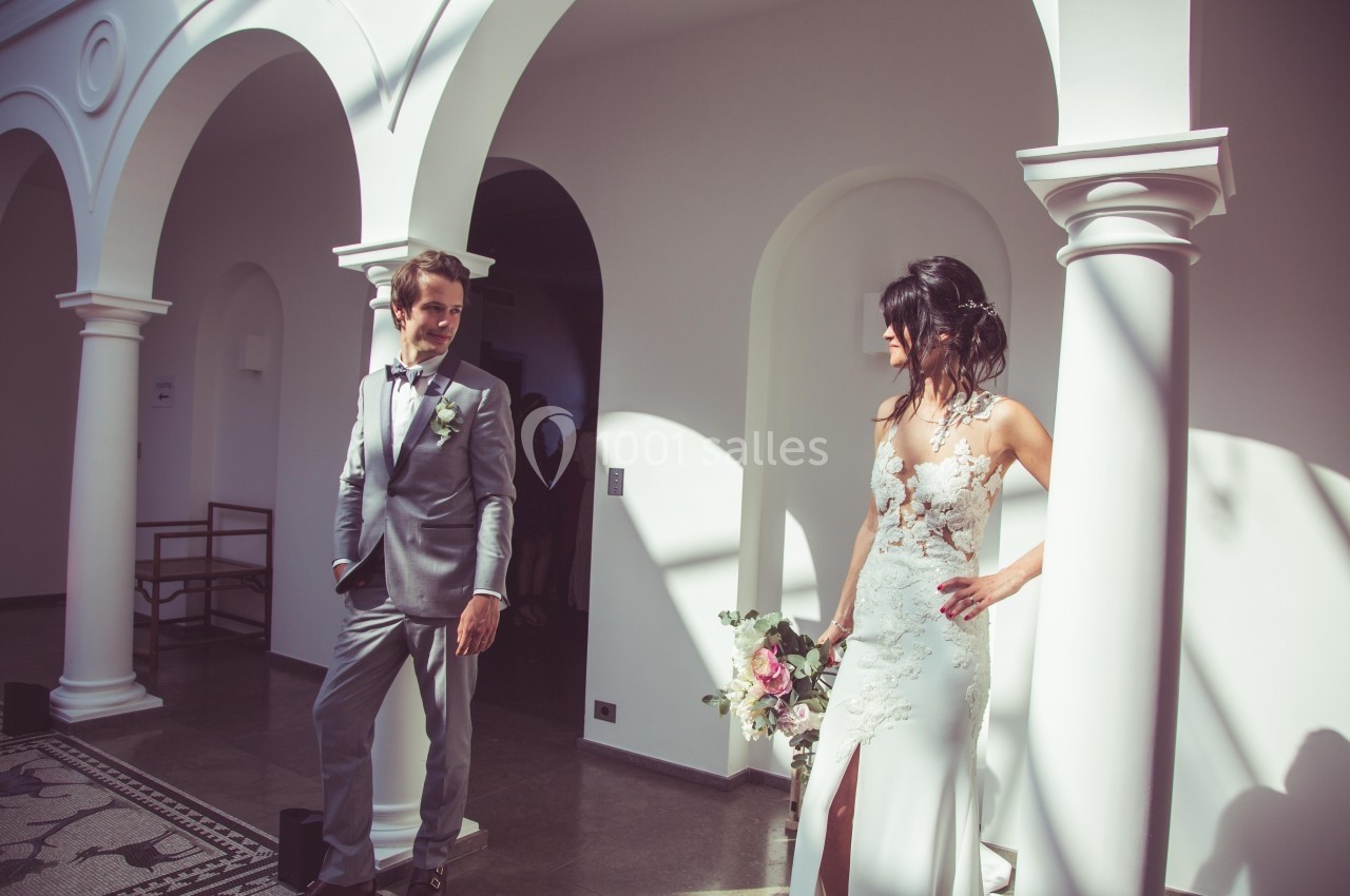 Un couple en tenue de mariage se regarde dans un bâtiment lumineux avec des arches blanches.