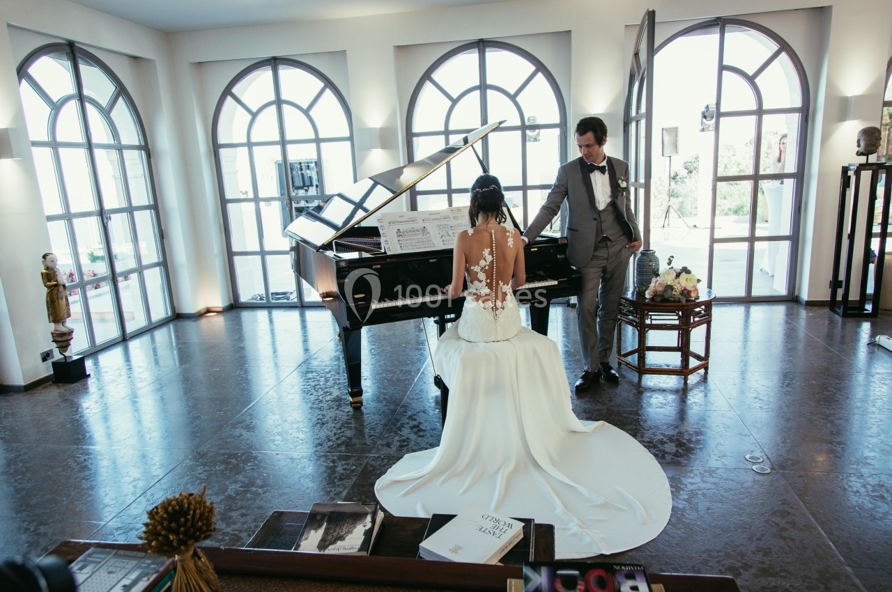 Une mariée en robe blanche joue du piano à queue, un homme en costume se tient à ses côtés dans une pièce lumineuse.
