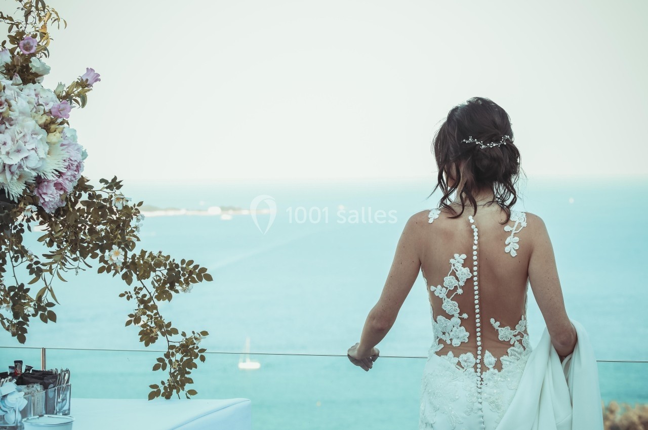 Une femme en robe de mariée dos nu regarde la mer depuis une terrasse, avec des fleurs à gauche.