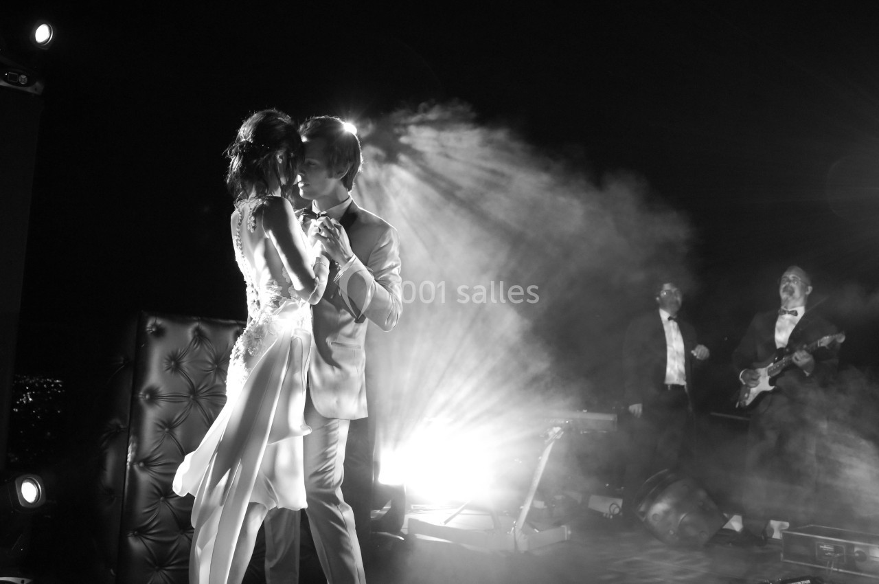 Un couple danse sous des projecteurs, entouré de fumée, avec des musiciens jouant en arrière-plan.
