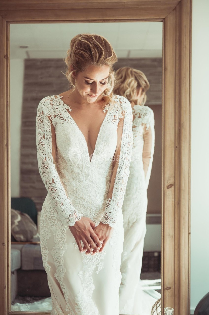Femme en robe de mariée en dentelle blanche, debout devant un miroir en bois dans une pièce lumineuse.