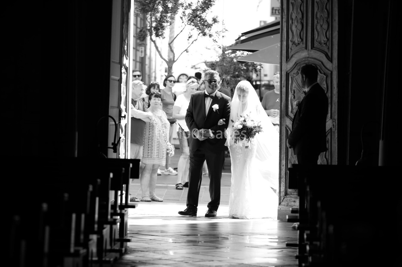 Un homme accompagne une mariée en robe blanche entrant dans une église, observés par des invités à l'extérieur.