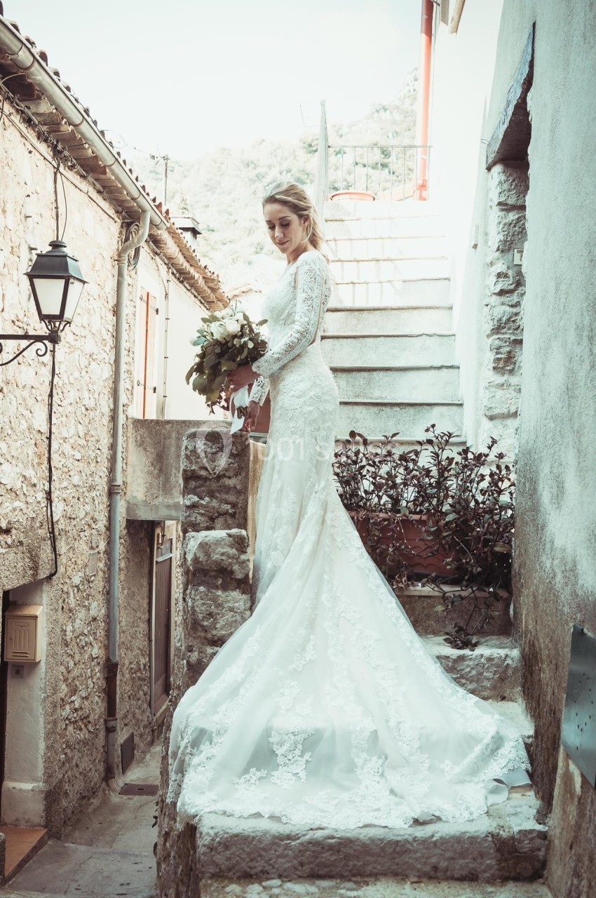 Femme en robe de mariée tenant un bouquet, posant dans une ruelle en pierre avec des escaliers en arrière-plan.
