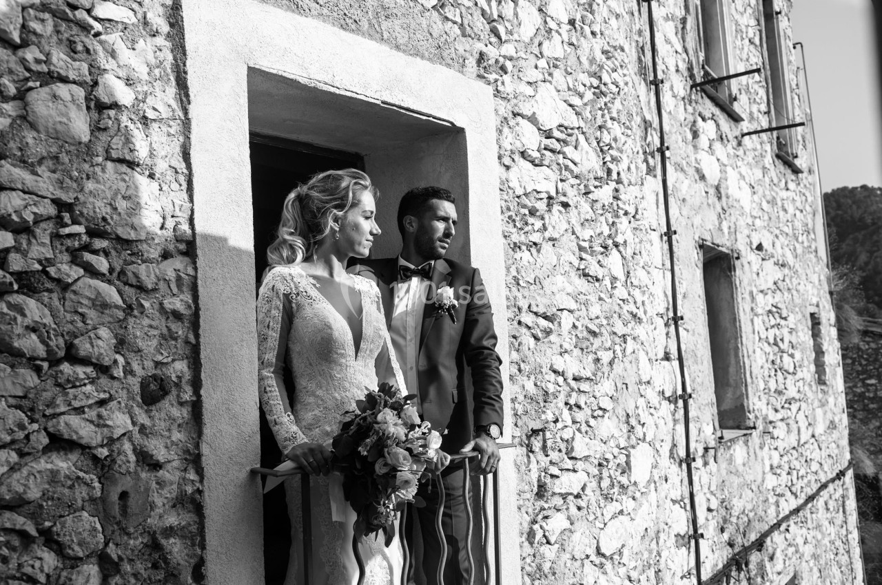 Un couple en tenue de mariage se tient sur un balcon en pierre, regardant au loin.