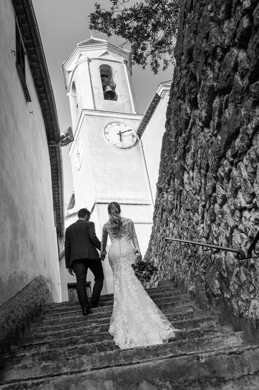 Un couple en tenue de mariage monte un escalier en pierre vers une église avec un clocher visible en arrière-plan.