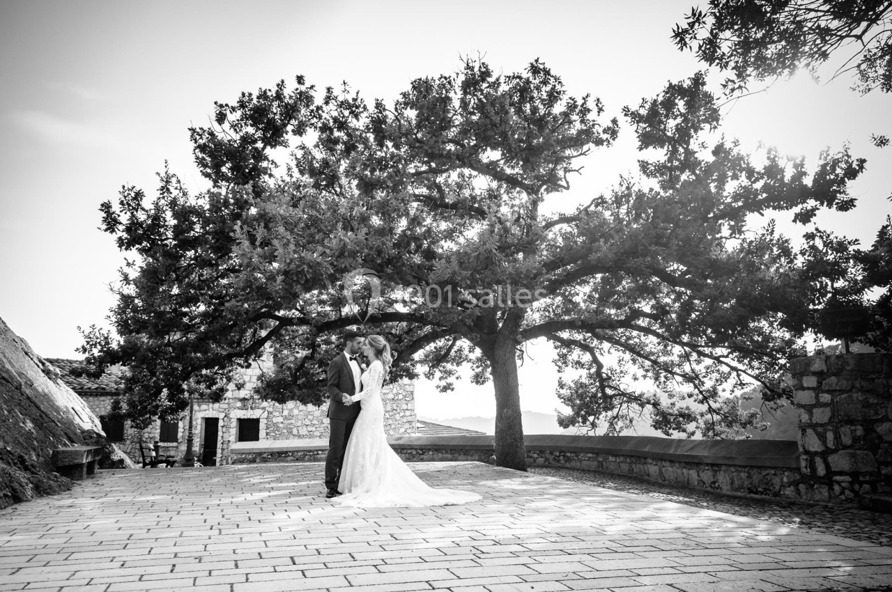 Un couple en tenue de mariage se tient sous un grand arbre sur une terrasse pavée, avec un paysage en arrière-plan.