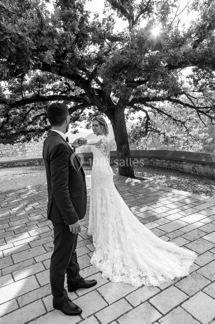 Un couple en tenue de mariage échange un regard sous un grand arbre, sur une terrasse pavée en extérieur.