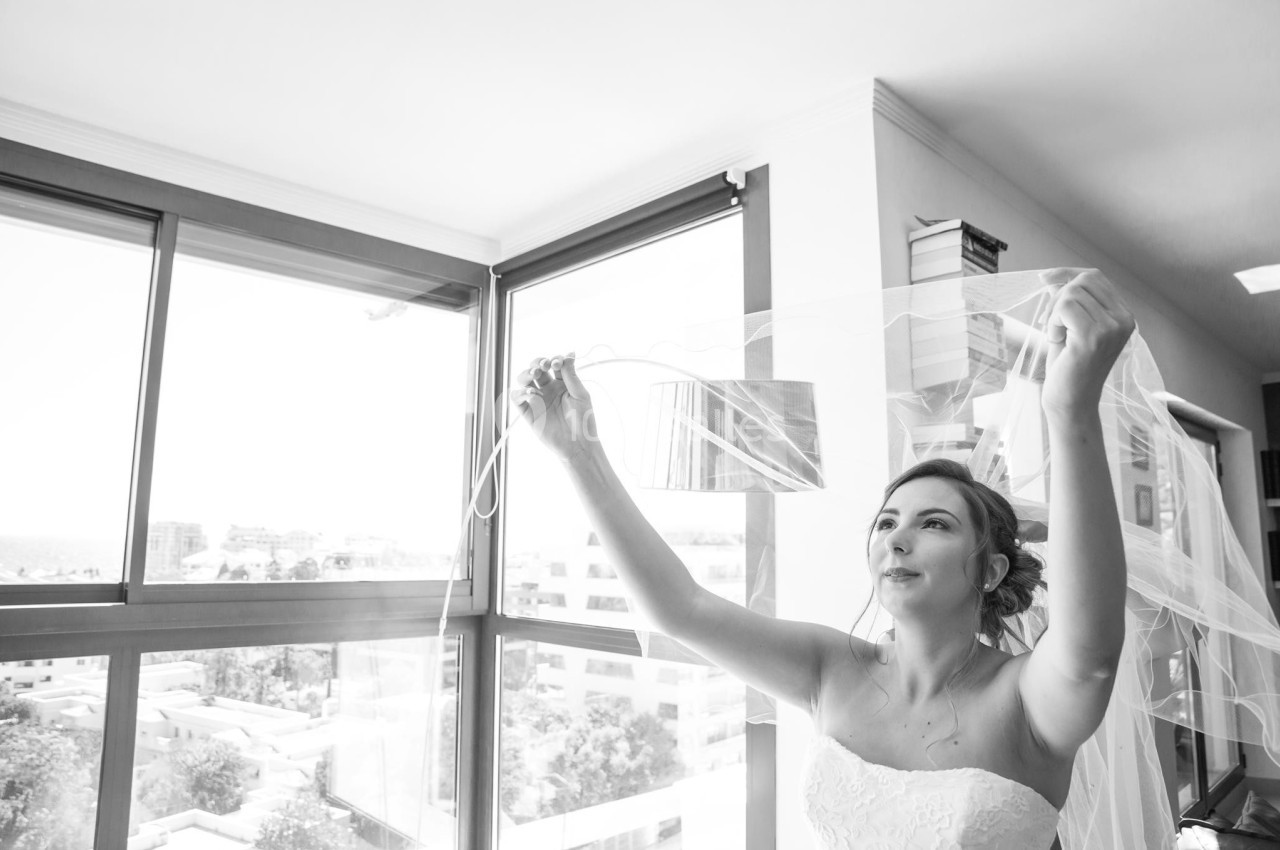 Une femme en robe de mariée ajuste son voile près d'une grande fenêtre laissant entrer la lumière naturelle.