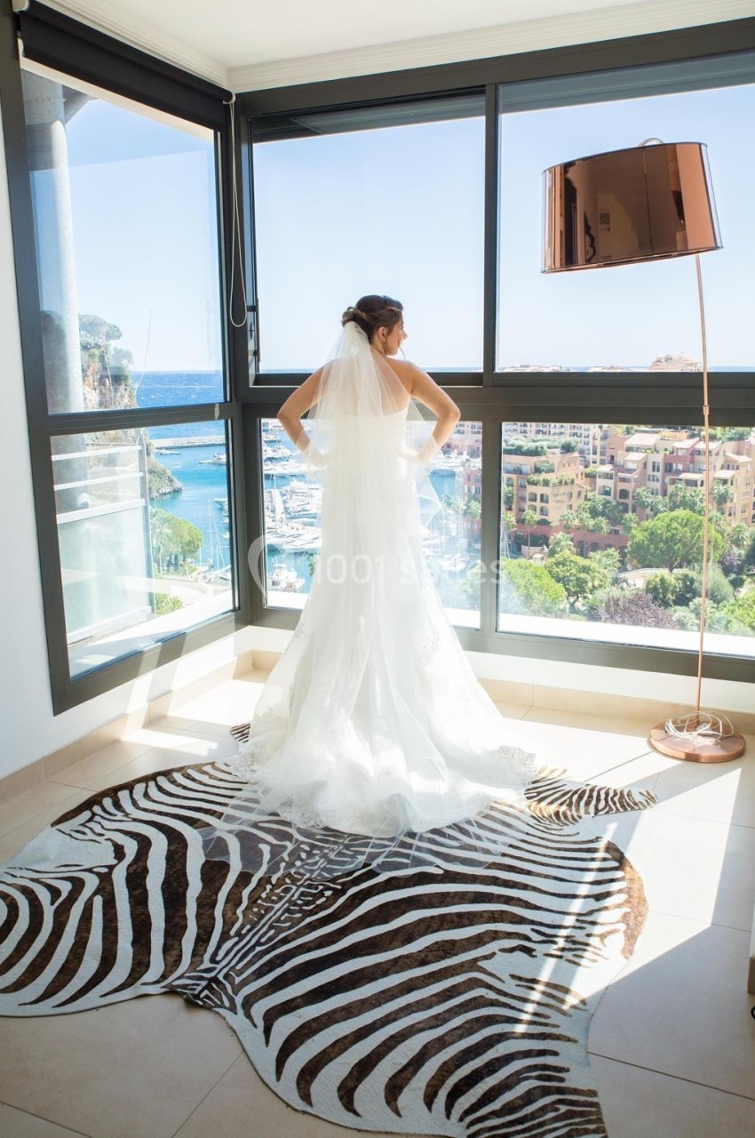 Une mariée en robe blanche regarde par une baie vitrée offrant une vue sur un port et des bâtiments ensoleillés.