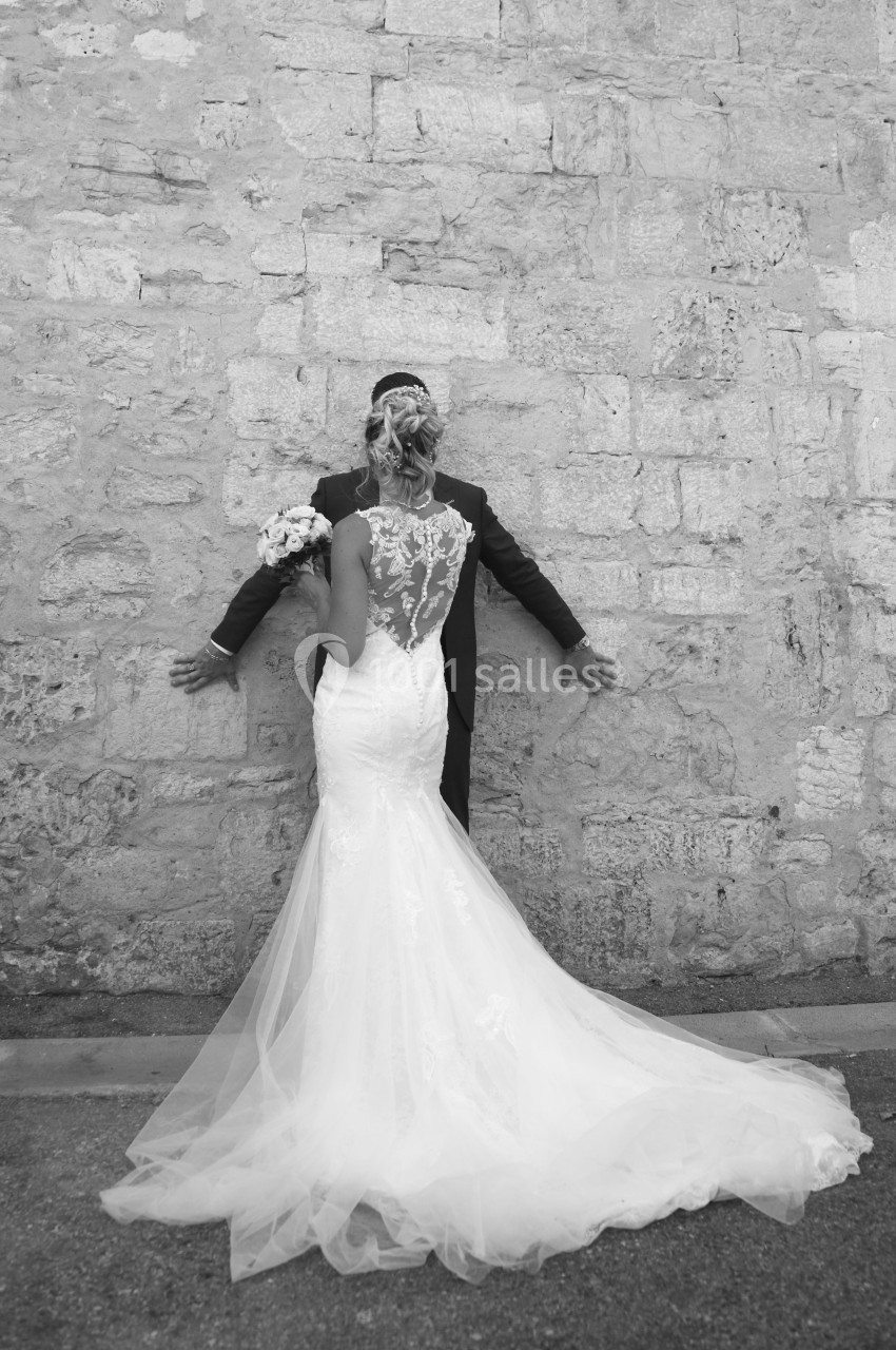 Une mariée en robe blanche et un homme en costume posent dos à un mur en pierre.
