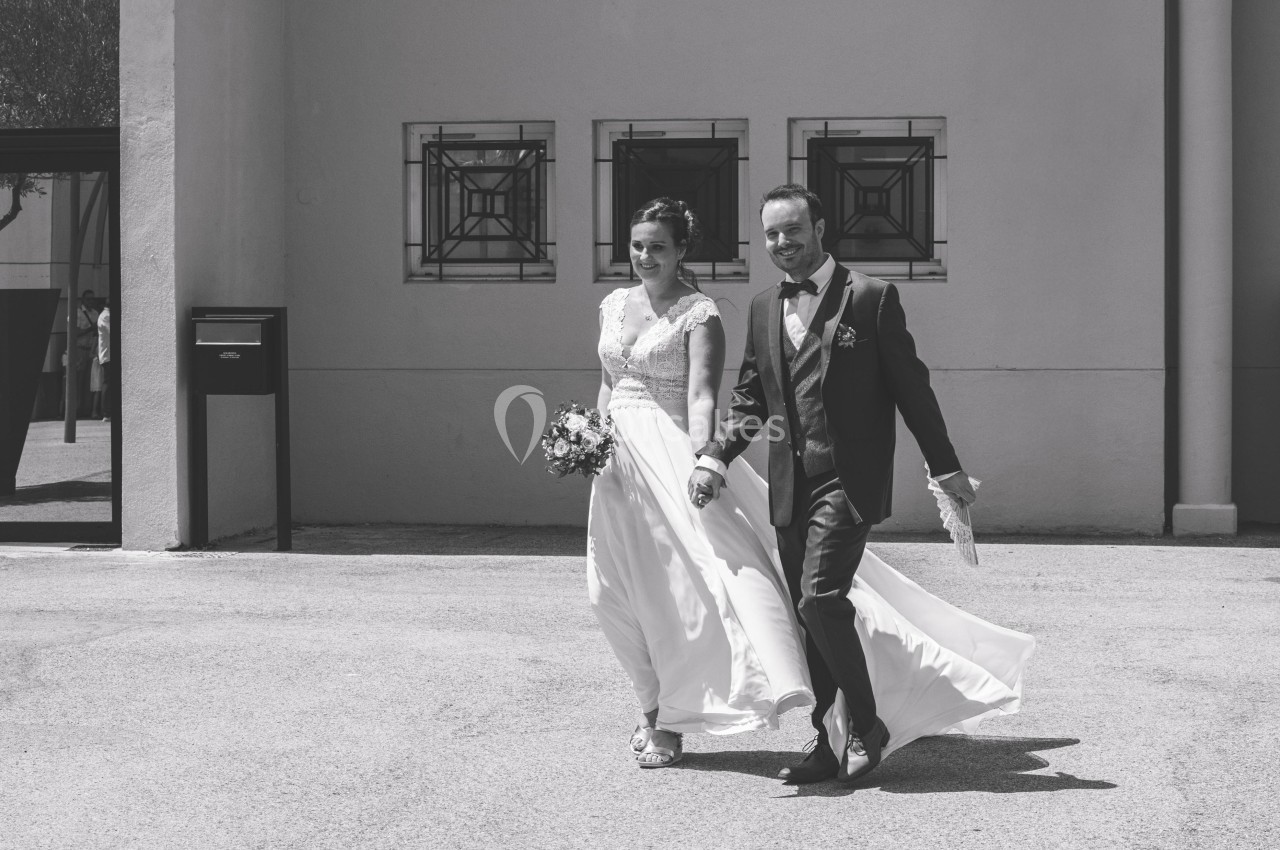 Un couple en tenue de mariage marche main dans la main devant un bâtiment clair.