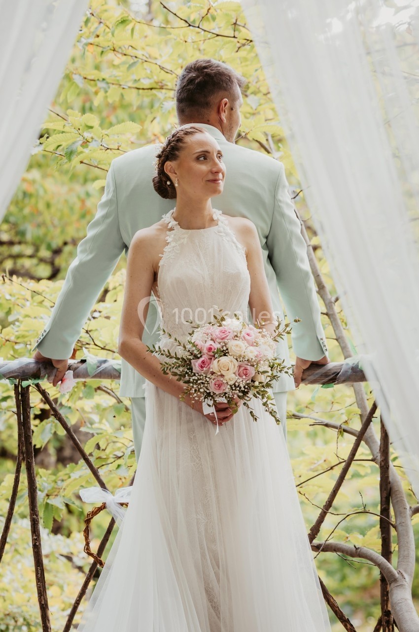 Un couple en tenue de mariage pose devant une balustrade en bois, entouré de verdure.