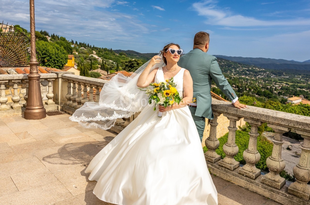 Une mariée souriante en robe blanche et lunettes de soleil tient un bouquet, avec un paysage vallonné en arrière-plan.