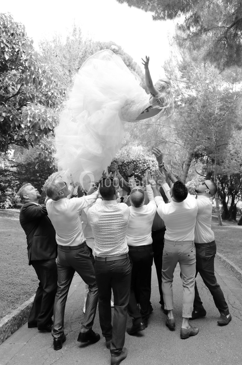 Des invités lancent une mariée en robe blanche en l'air lors d'une célébration en extérieur.