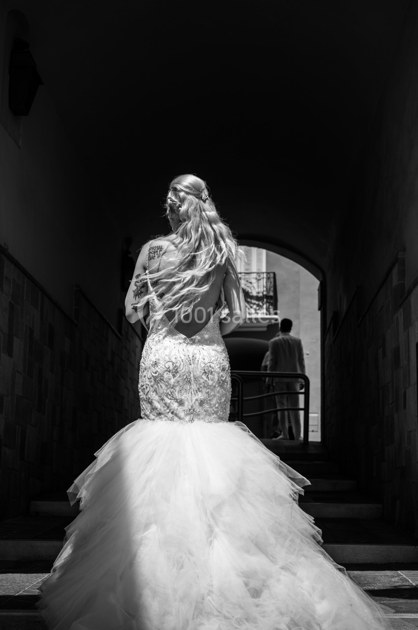 Une femme en robe de mariée dos nu monte des escaliers dans un passage sombre, éclairée par une lumière naturelle.
