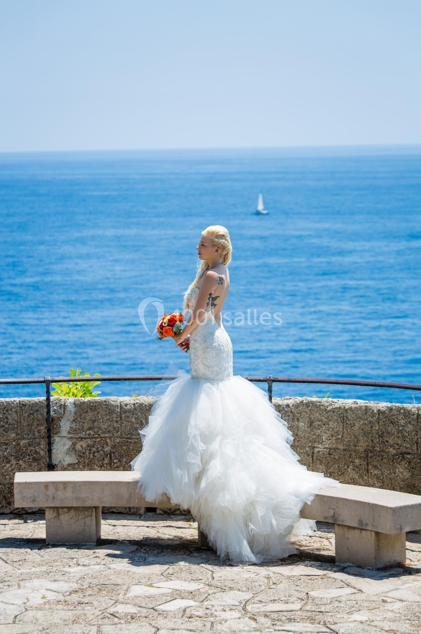 Femme en robe de mariée blanche tenant un bouquet, debout près d'un muret avec vue sur la mer et un voilier au loin.