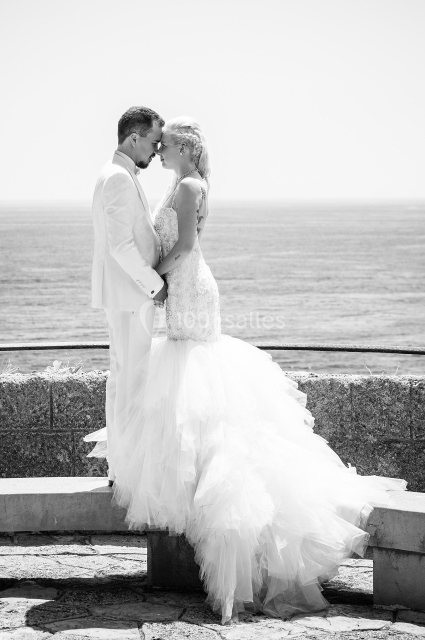 Un couple en tenue de mariage s'embrasse devant une vue dégagée sur la mer.