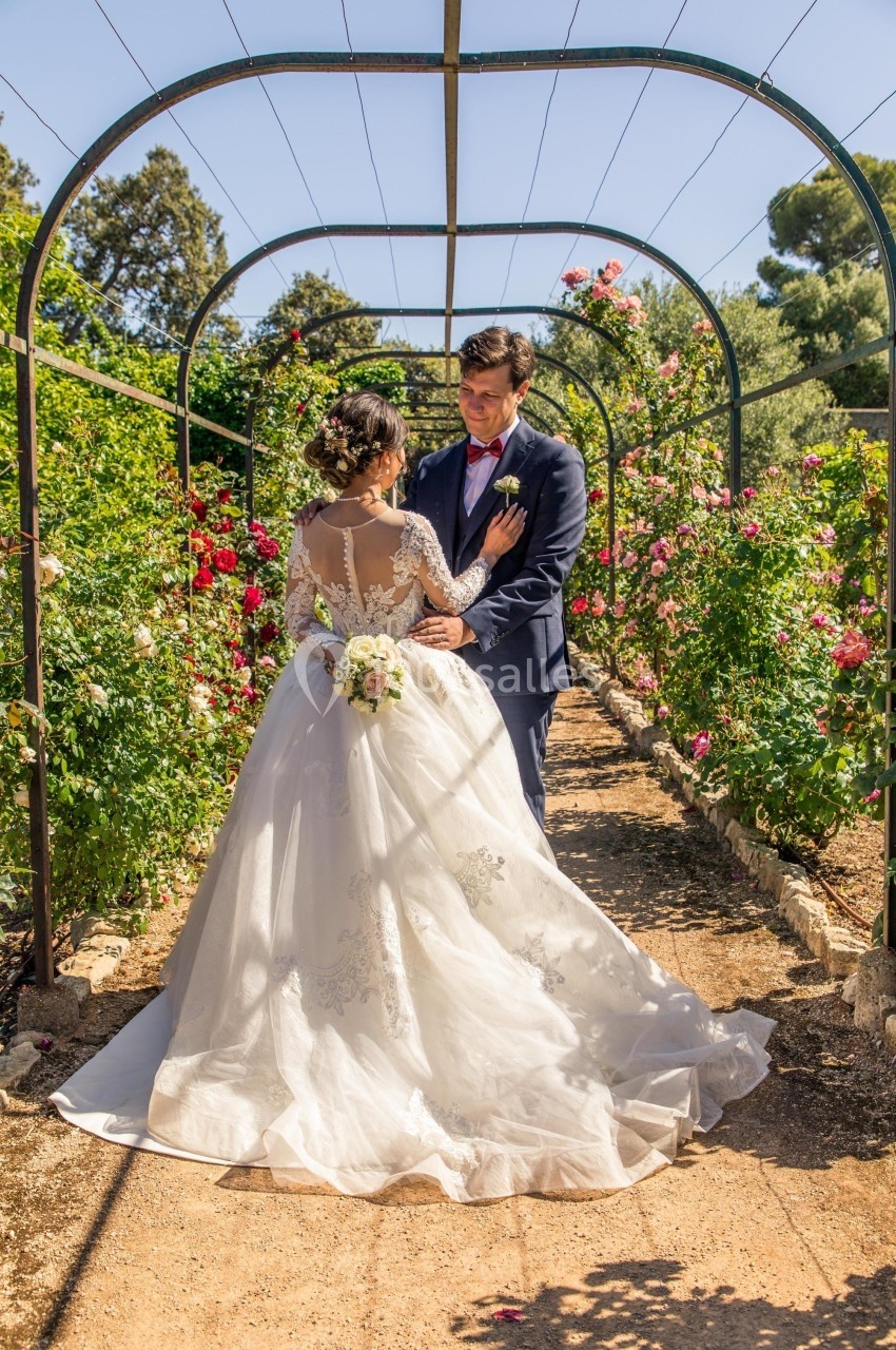 Un couple en tenue de mariage se tient sous une arche fleurie dans un jardin ensoleillé.