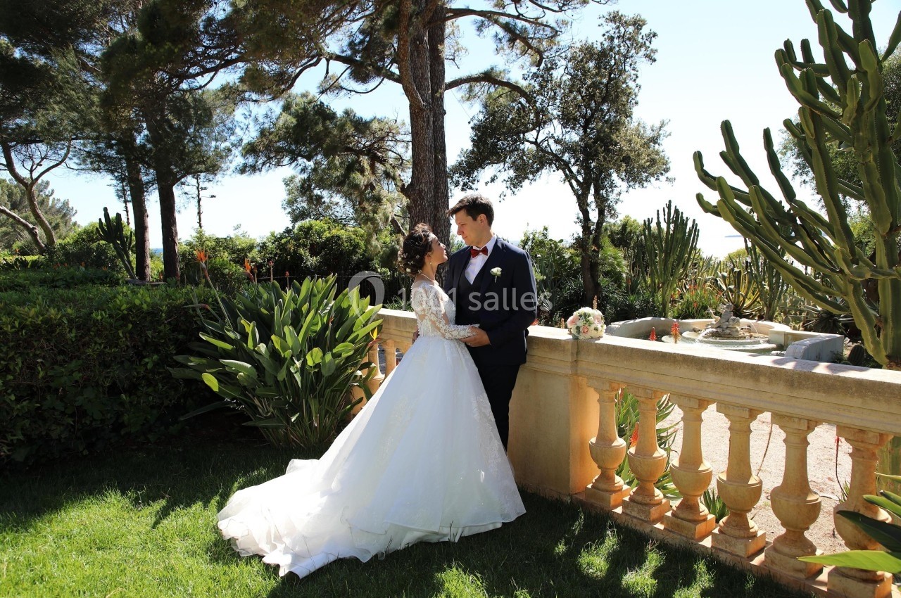 Un couple en tenue de mariage se tient près d'une balustrade dans un jardin verdoyant sous un ciel dégagé.