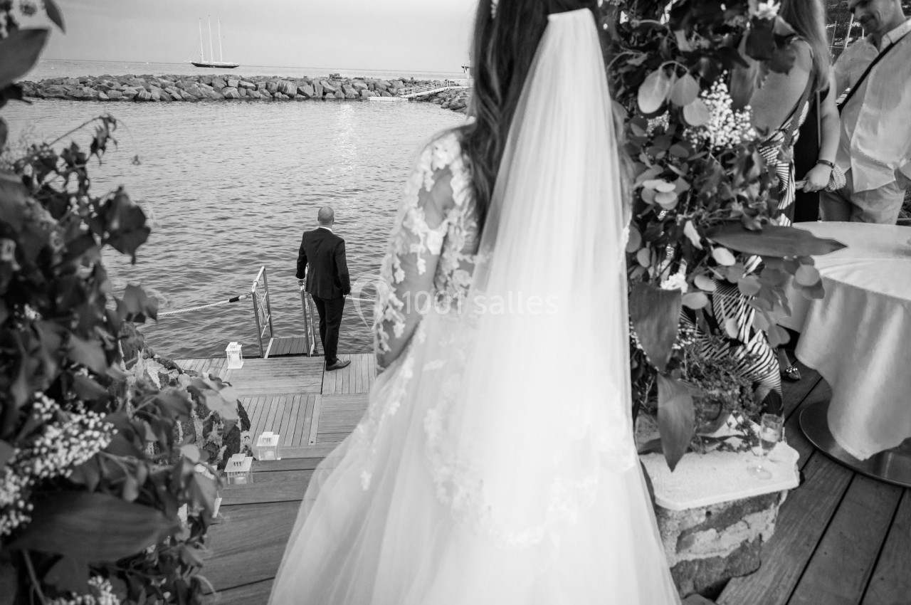 Une mariée en robe blanche observe un homme en costume marchant vers un quai au bord de l'eau.