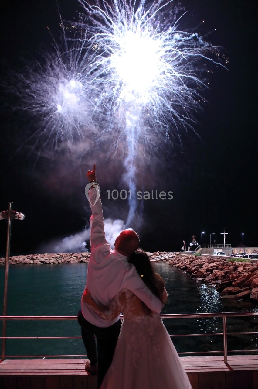 Un couple de dos admire un feu d'artifice éclatant au-dessus d'une jetée bordée d'eau la nuit.