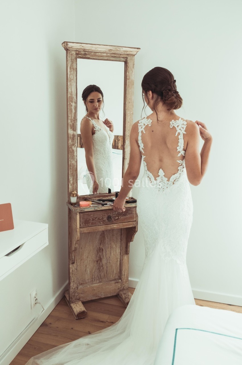 Femme en robe de mariée ajustant sa tenue devant un miroir ancien dans une pièce lumineuse.