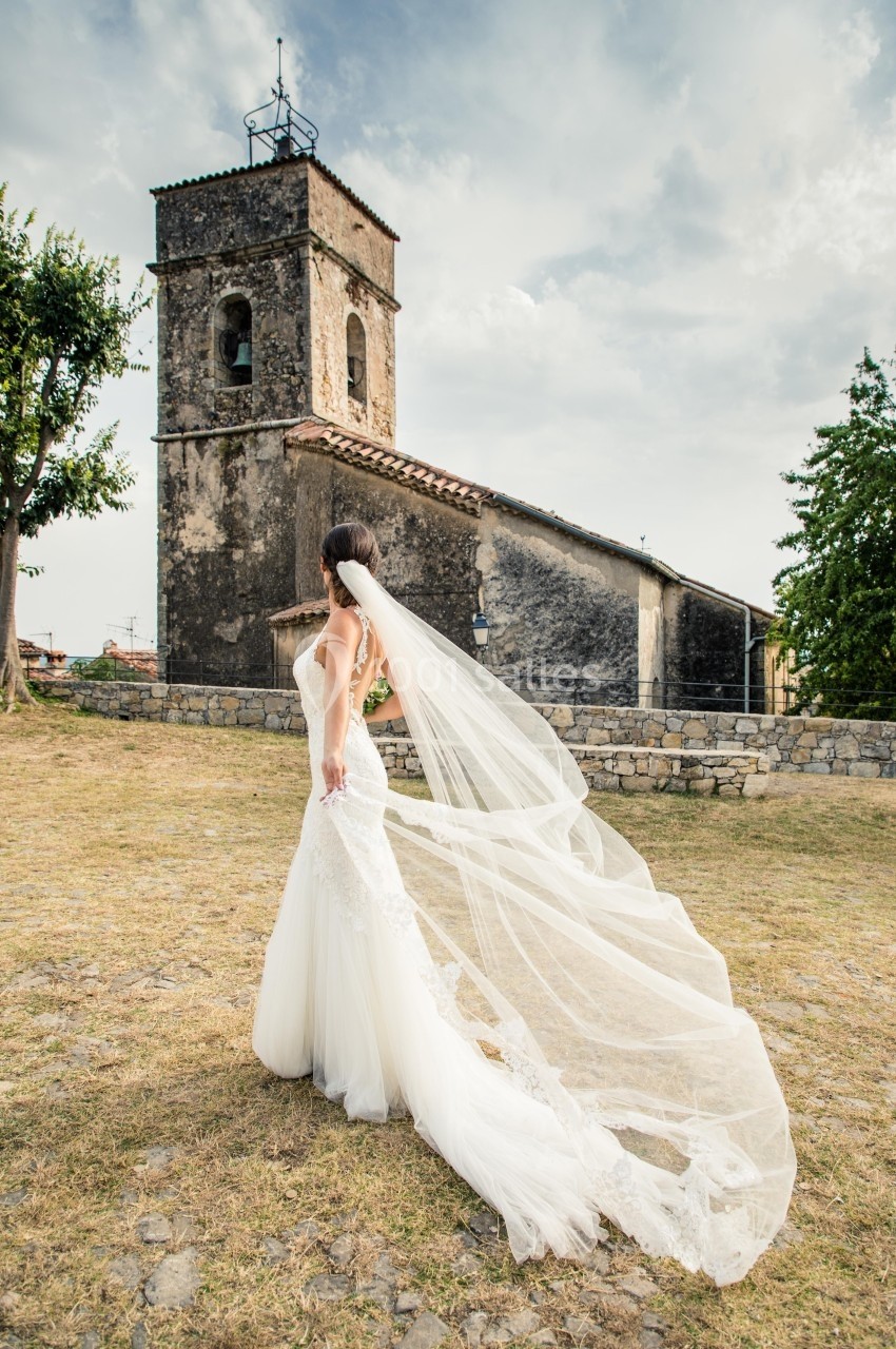 Femme en robe de mariée avec un long voile, debout devant une église en pierre dans un paysage rural.