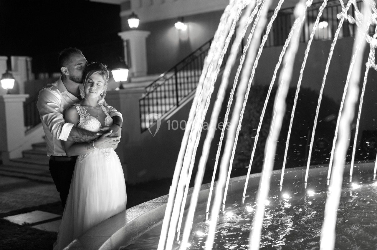 Un couple en tenue de mariage s'enlace près d'une fontaine illuminée, de nuit.