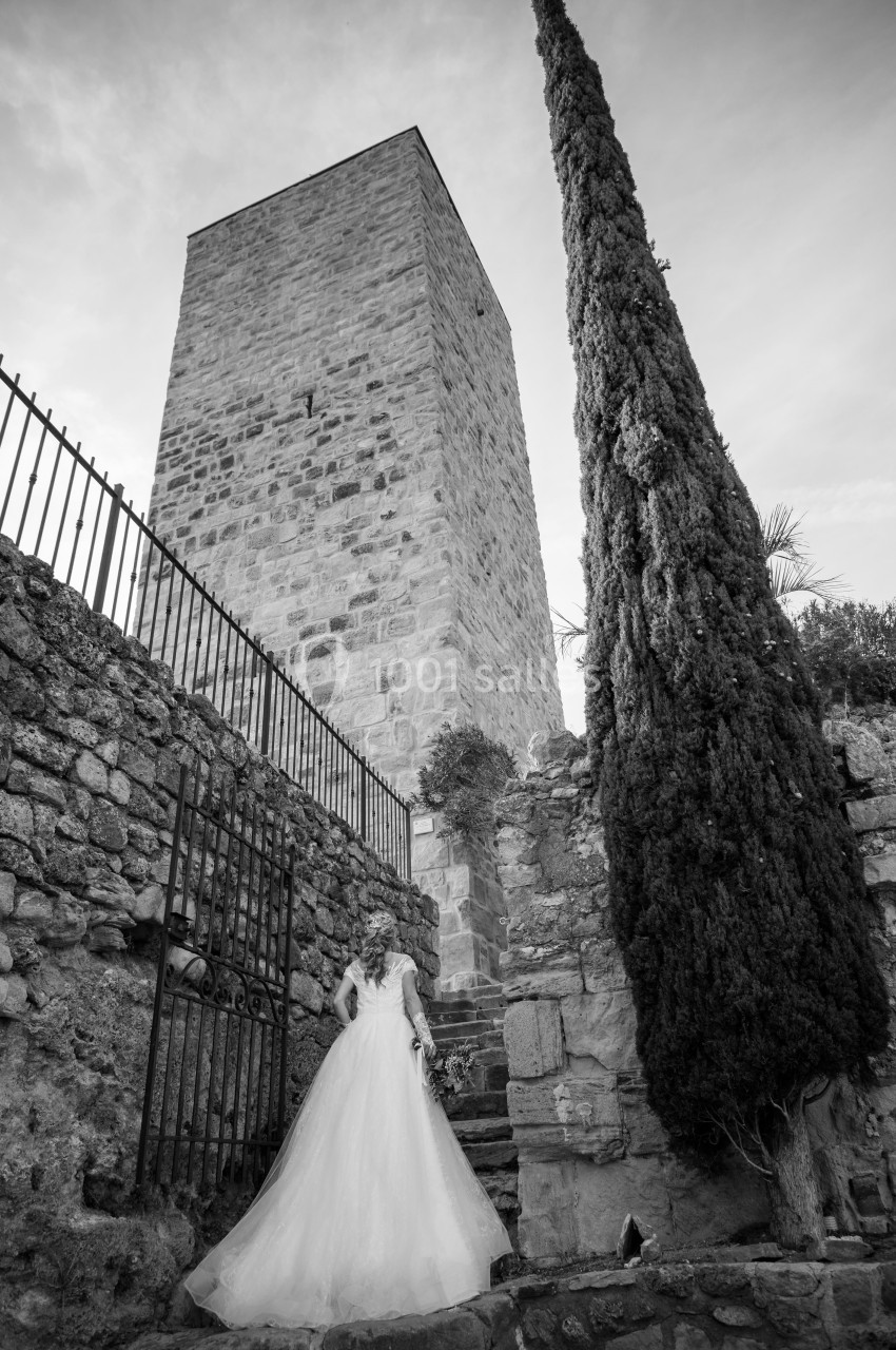 Une mariée en robe blanche monte des marches en pierre devant une tour médiévale et un cyprès.