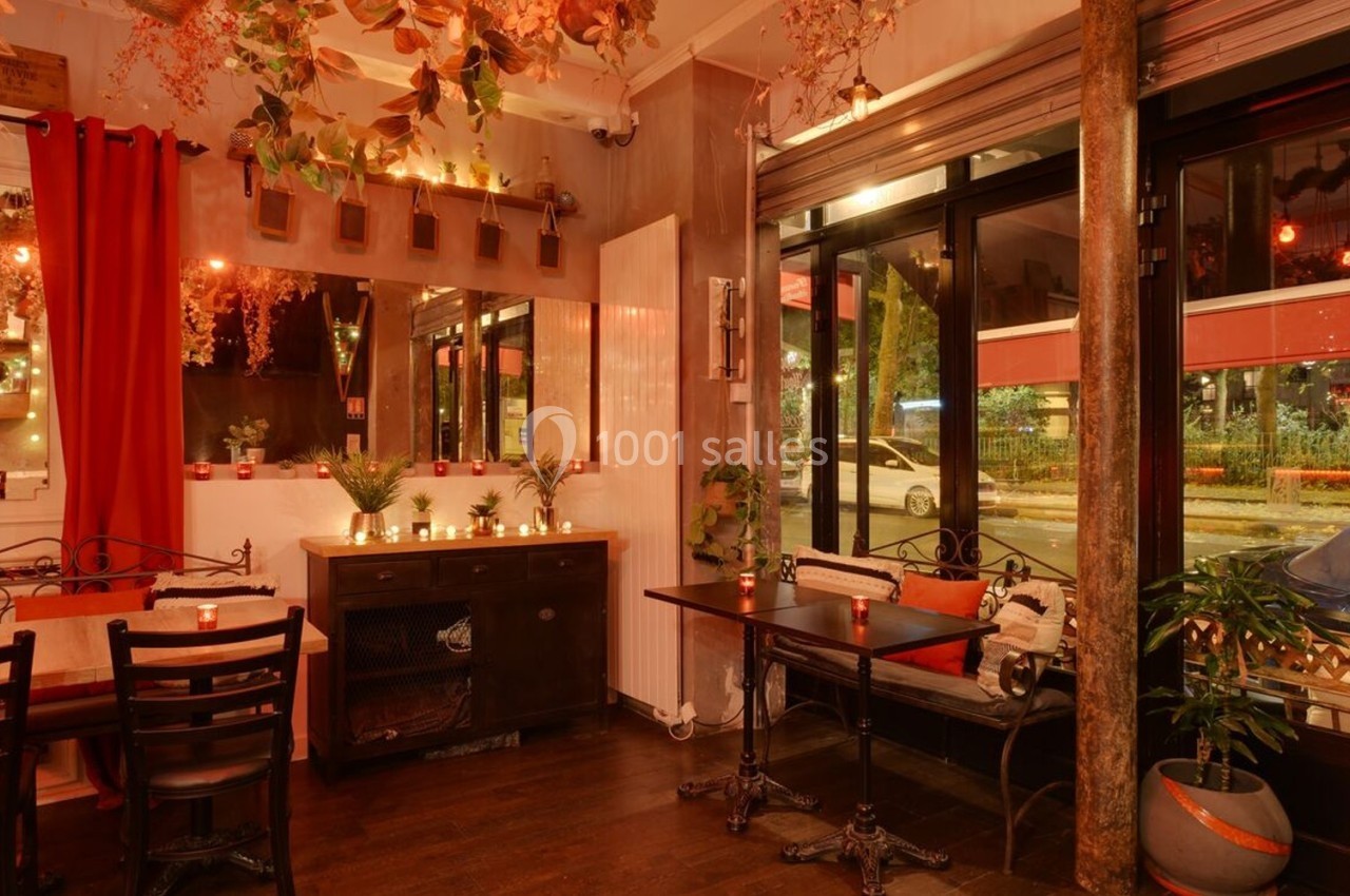 Salle de restaurant chaleureuse avec décoration végétale, éclairage tamisé et grandes baies vitrées donnant sur la rue.