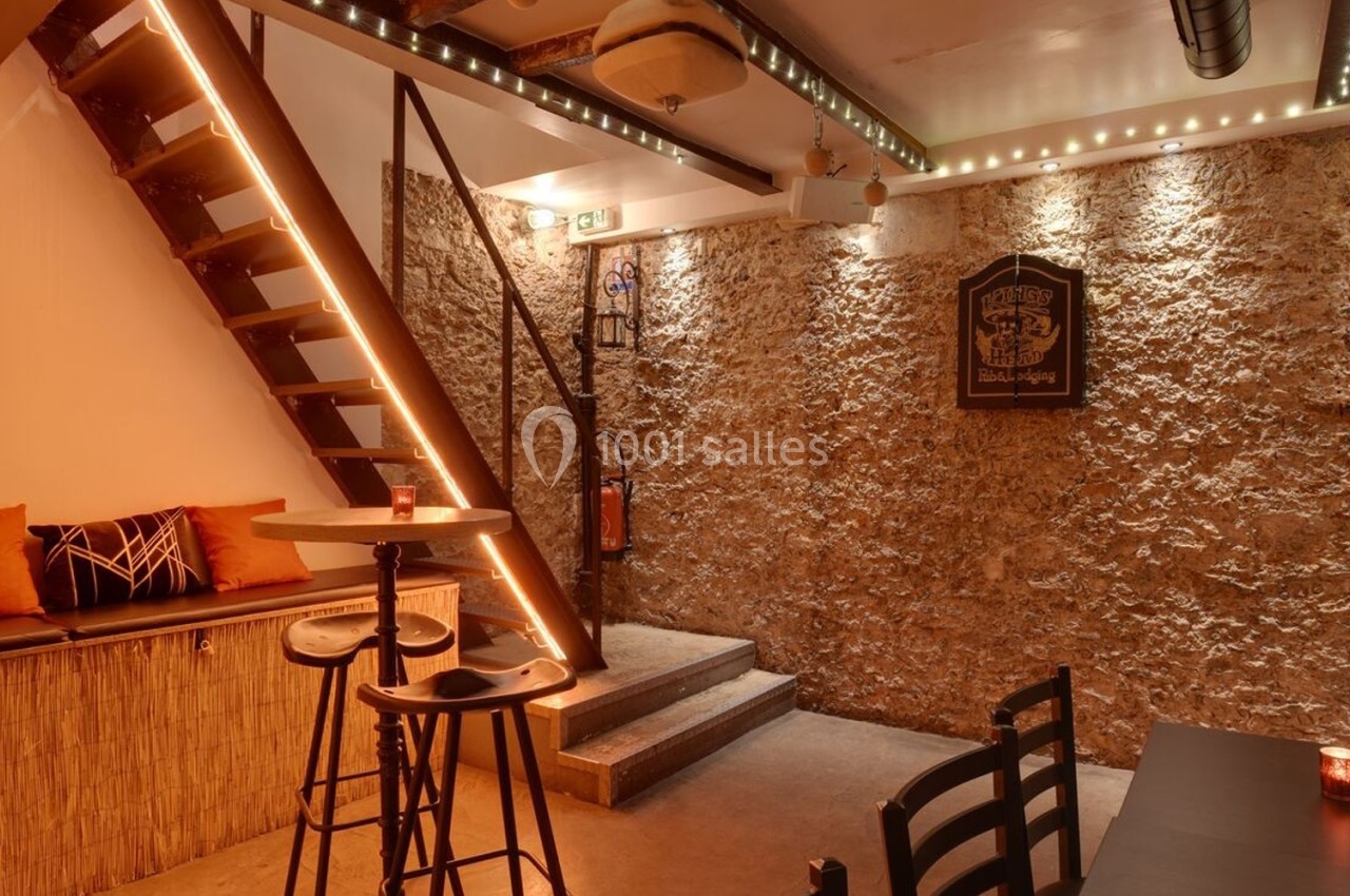 Espace intérieur chaleureux avec murs en pierre, escalier en bois, table haute et éclairage tamisé.