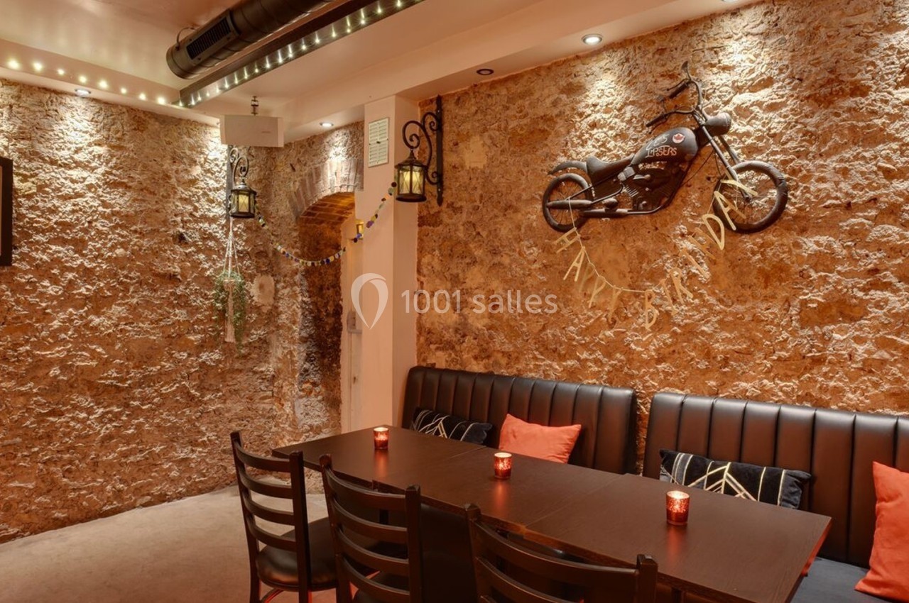 Salle chaleureuse avec murs en pierre, table en bois, banquette noire, décoration murale et éclairage tamisé.