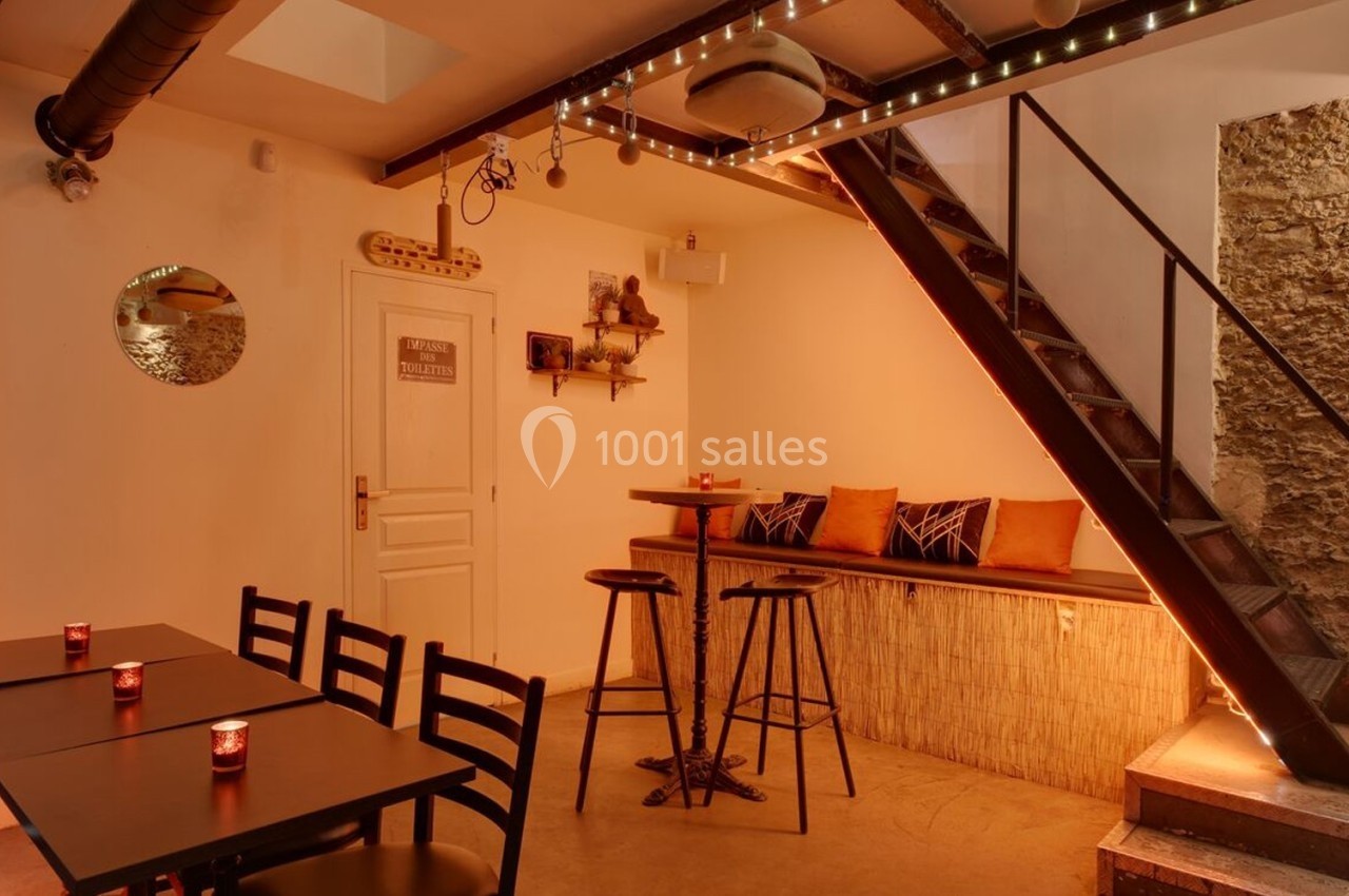 Salle chaleureuse avec tables, chaises, banquette, escalier métallique et éclairage tamisé.