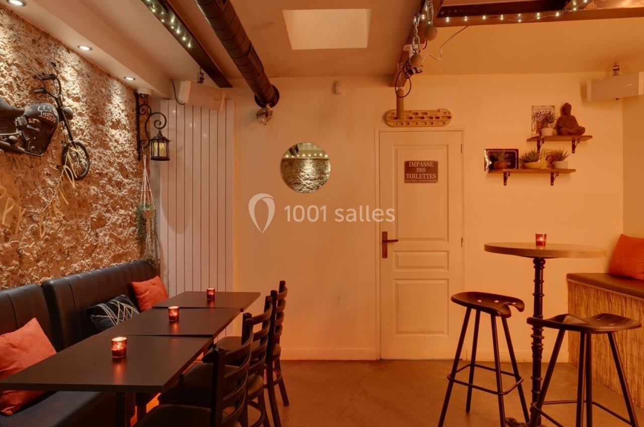 Salle chaleureuse avec murs en pierre, tables et chaises noires, coussins orange, et éclairage tamisé.